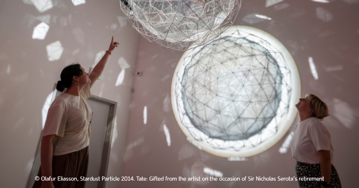 Two women stood underneath a white circular light artwork installation named Stardust Particle.